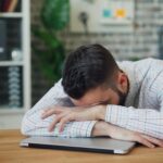 Tired office worker resting head on laptop at desk in a modern office.