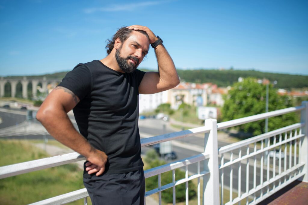 Bearded man in black t-shirt standing on a footbridge in Portugal's urban landscape on a sunny day.