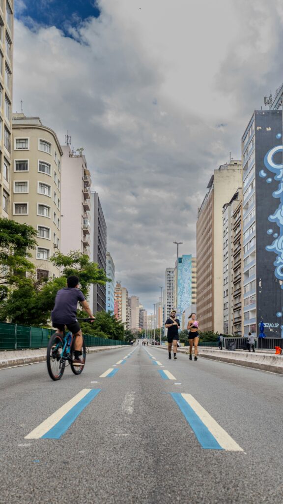 A lively street in São Paulo, Brazil, with people cycling and jogging under a clouded blue sky.